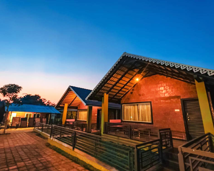 Side view of cozy cottages at dusk with warm lighting. The cottages have slanted roofs and are made of brick with large windows. They are surrounded by a paved area and simple metal railings, with trees and a clear evening sky in the background, offering a glimpse of Dandeli nature.