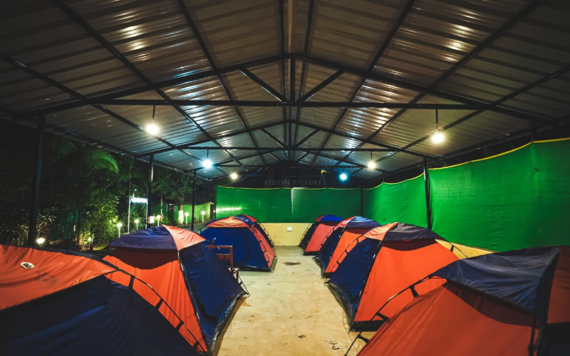 A covered outdoor area illuminated by hanging lights, with rows of colorful camping tents placed neatly on the ground. The area is surrounded by green mesh walls and some foliage is visible in the background. The setting appears organized and inviting for campers seeking a Dandeli adventure.