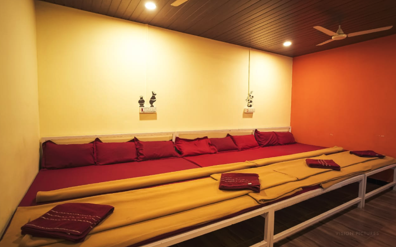 A cozy room with a long cushioned seating area adorned with red and yellow cushions. The ceiling is wooden with recessed lighting and a ceiling fan. The walls are painted in warm shades of orange and cream, decorated with small wall ornaments above the seats, evoking the charm of Dandeli resorts.