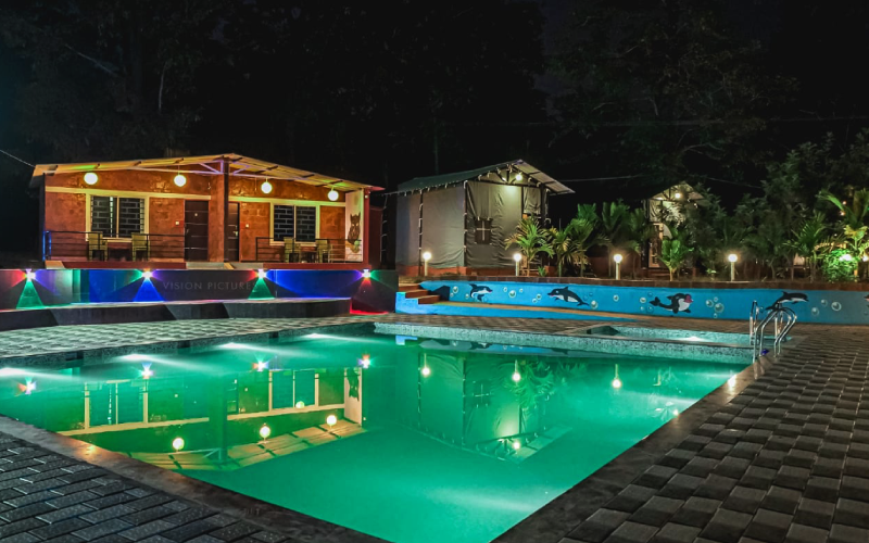 A night view of a swimming pool illuminated with underwater lights. Adjacent to the pool are wooden cabins with terraces, also lit up. The pool area is decorated with multicolored lights, and a garden with trees and plants surrounds the area, creating a serene atmosphere reminiscent of a Dandeli home stay.