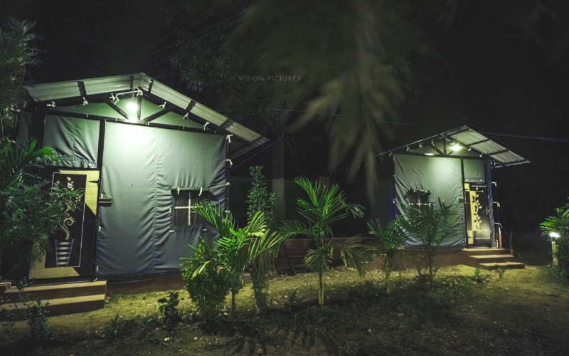 A nighttime scene featuring two small, tent-like structures with illuminated entrances at a Dandeli home stay. The structures are surrounded by lush greenery, including various small plants and trees. The ground appears to be bare soil, creating a serene and calm ambiance.