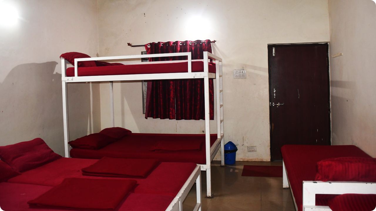 A small, simply furnished room with a bunk bed and a single bed, all featuring red bedding. The walls are white, and there's a window with red curtains. A closed wooden door is on the right side. The room is lit by a single overhead light, perfect for those looking for Dandeli Resort and homestay Packages.