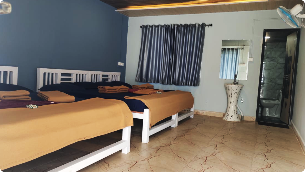 A well-kept hotel room at Dandeli Resort and Homestay Packages features two white wooden beds covered in dark blue bedsheets and orange blankets, with folded towels placed on top. The room also includes a window with closed grey curtains, a sink with a mirror, and a private bathroom in the corner.