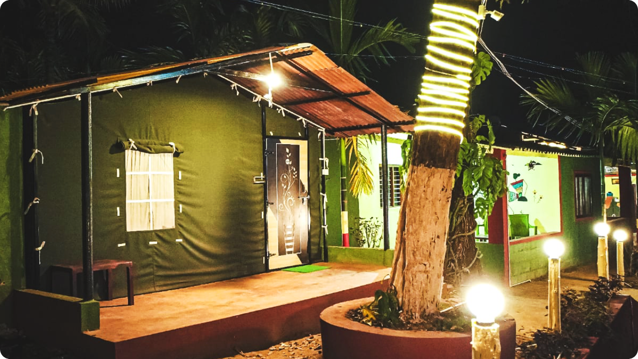 A cozy, green cottage with a brightly lit exterior sits amidst palm trees in an exclusive resort and homestay packages in Dandeli. The porch area has a single chair, and the building is adorned with string lights. A tree wrapped in yellow lights stands in front of the cottage, with pathway lights on either side.