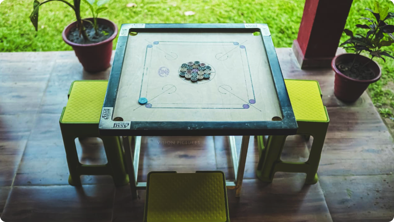 A carrom board with arranged pieces in the center is placed on a square table outdoors at Dandeli Resort and Homestay Packages. The table has four green stools on each side, with two potted plants nearby on a wooden floor and a grassy area visible in the background.