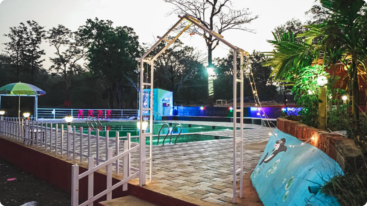 A well-lit outdoor swimming pool area at dusk with poolside seating and umbrellas. A partially visible mural with dolphins is on the right side, and trees surround the area in the background. The entrance to this serene oasis at Dandeli Resort and Homestay Packages features a decorative white archway.