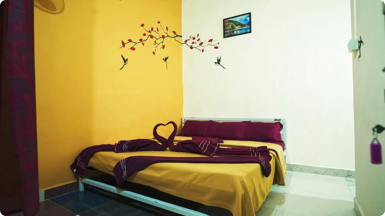 A cozy bedroom with a yellow and white wall invites relaxation. The bed features bright yellow bedding, dark red pillows, and a blanket folded creatively into a heart shape. A wall decal of birds and flowers adorns the yellow wall, with a framed picture above it, reminiscent of Dandeli Resort and Homestay Packages.