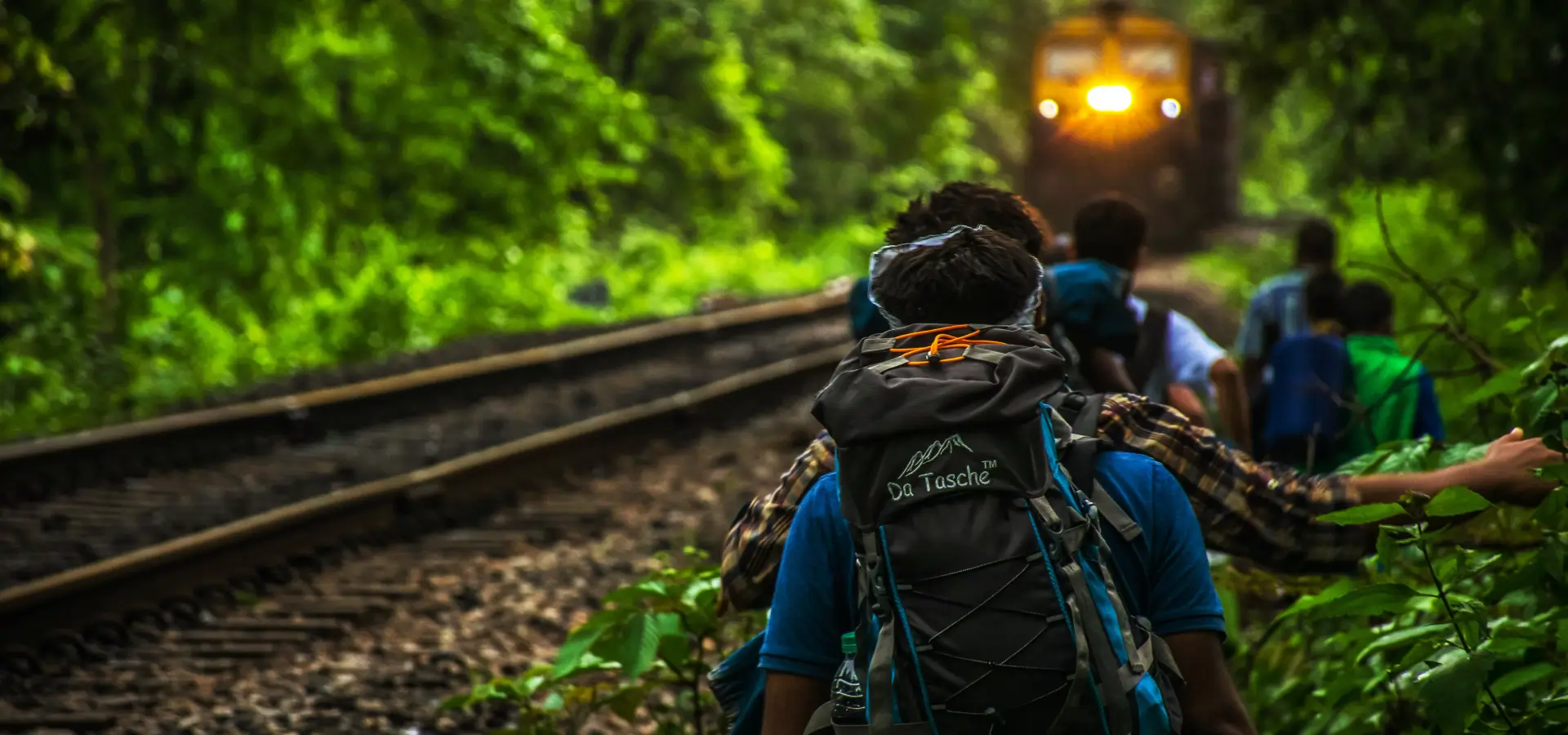 A group of hikers with backpacks walk parallel to train tracks in a lush, green forest. In the distance, a train with its headlight on approaches, creating a glowing effect amidst the verdant surroundings. The hikers appear to be cautious of the approaching train, appreciating Dandeli nature.