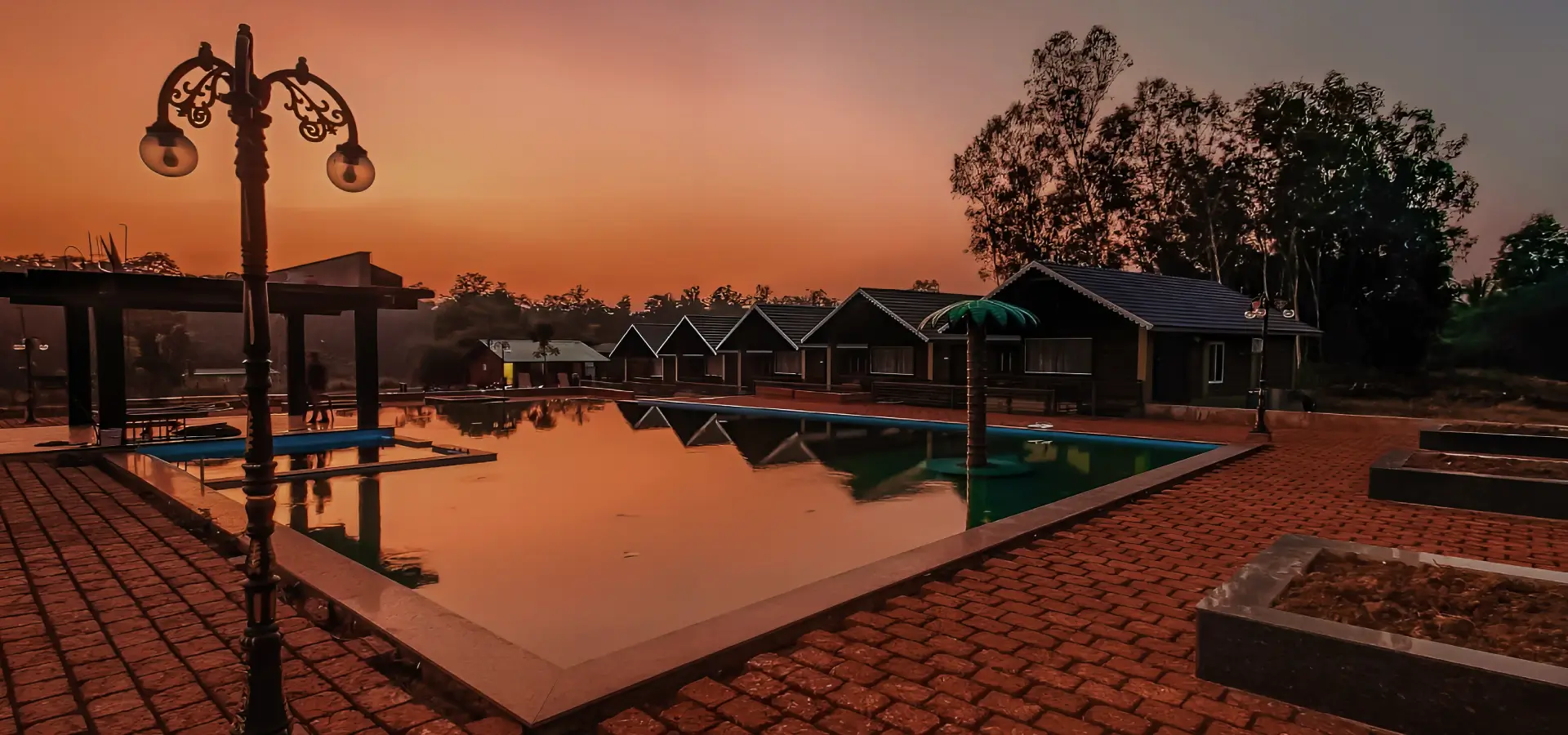 A serene evening view of Dandeli resorts featuring a large rectangular swimming pool. The pool is surrounded by brick-paved walkways and bordered by multiple identical cabin-like structures. The sky is a warm gradient from orange to red, and tall trees frame the background.